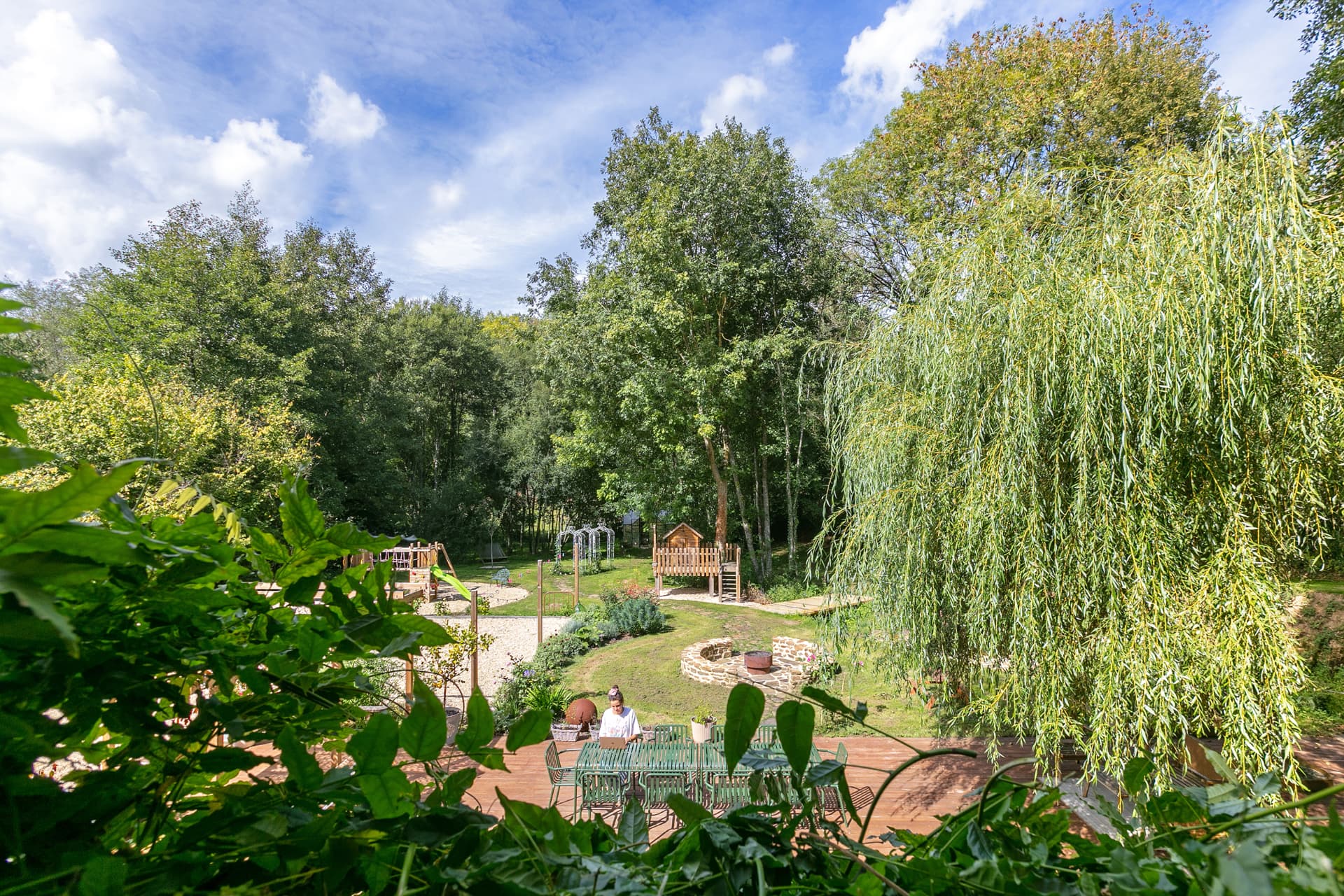 Garden deck under a weeping willow, with treehouse, fire pit and play area beyond